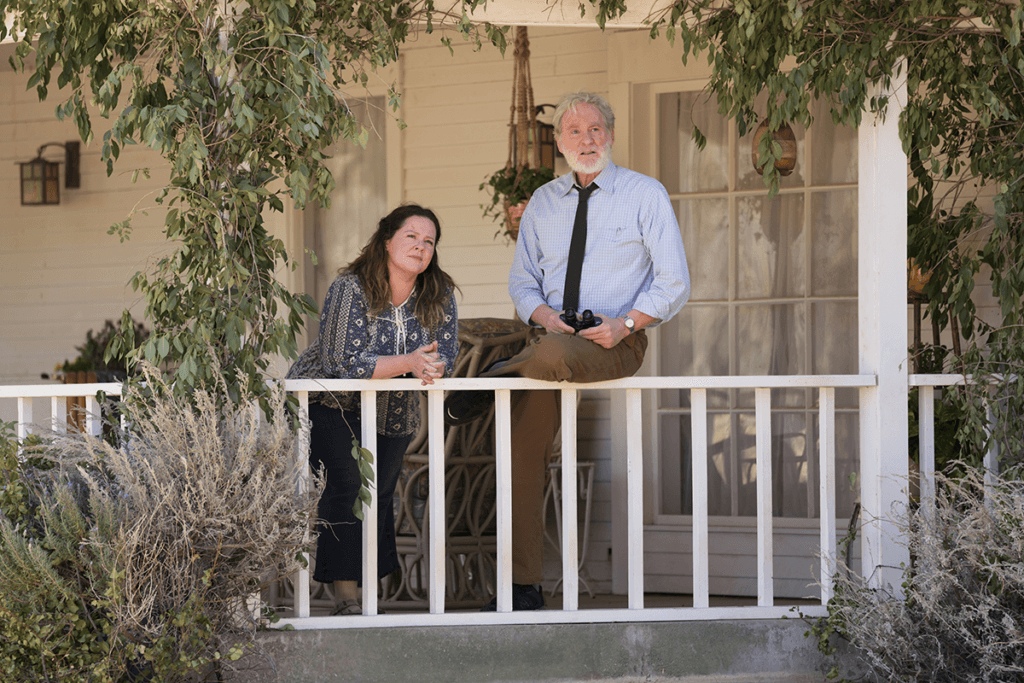 Melissa McCarthy e Kevin Kline lado a lado olhando para o horizonte na varanda. Ela encostada e ele sentado no parapeito