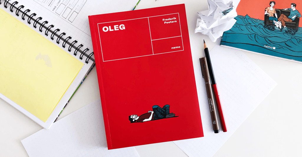 Quadrinho Oleg centralizado, envolta papeis, lapis, caneta