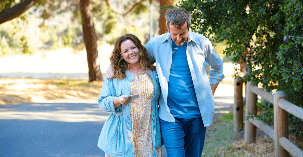 Um casal,  Melissa McCartthy e Chris O'Dowd abraçados enquento caminham em uma rua replata de árvores